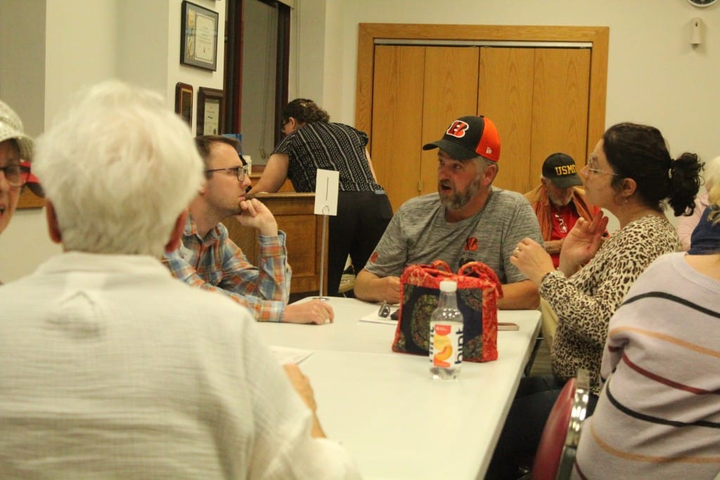 Several people sit around a table in conversation, with others seated and standing in the background.