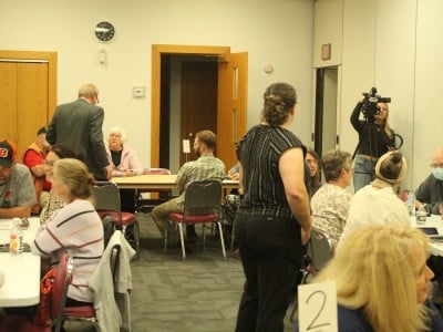 A crowded room, with several people sitting at three different tables and others standing.