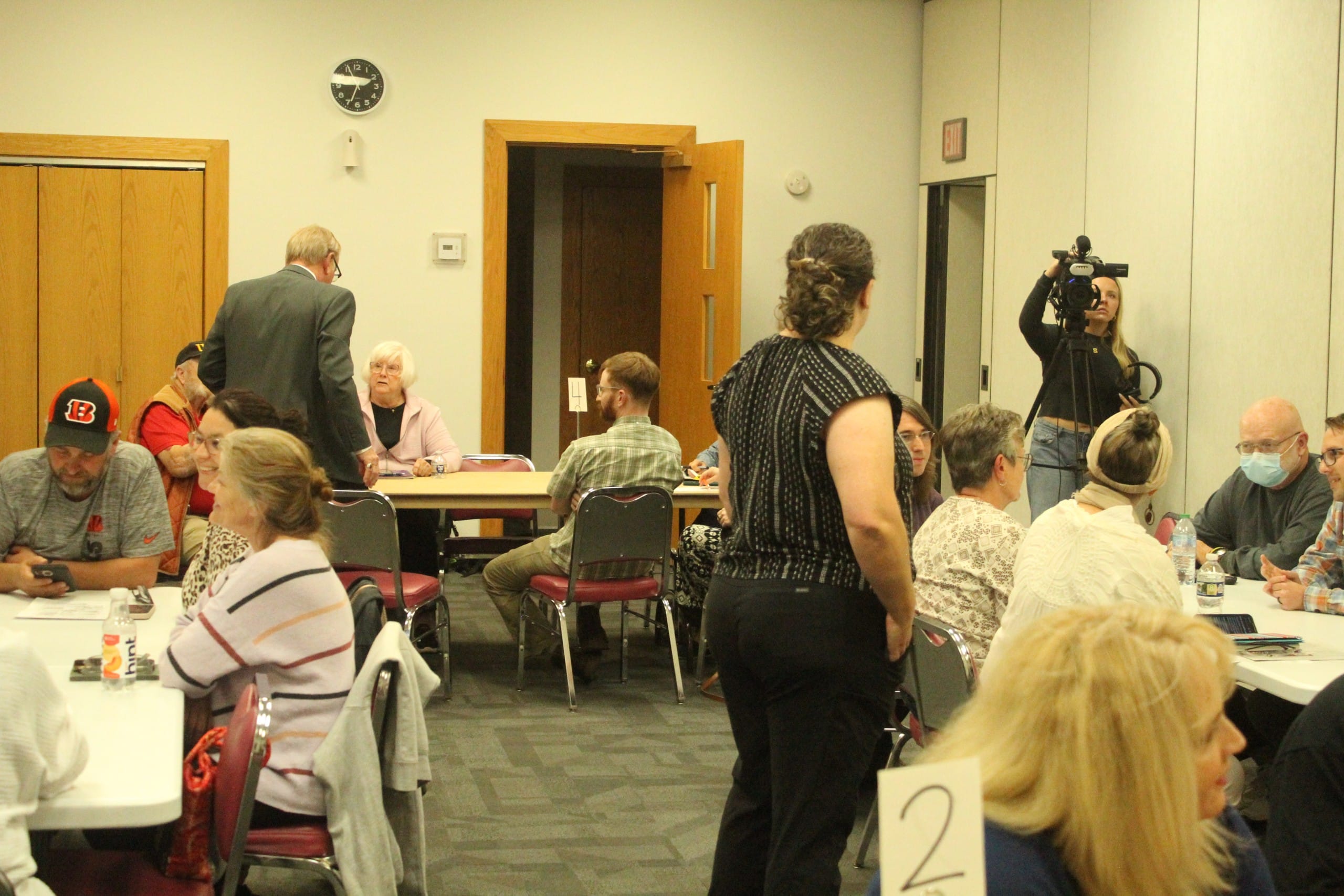 A crowded room, with several people sitting at three different tables and others standing.