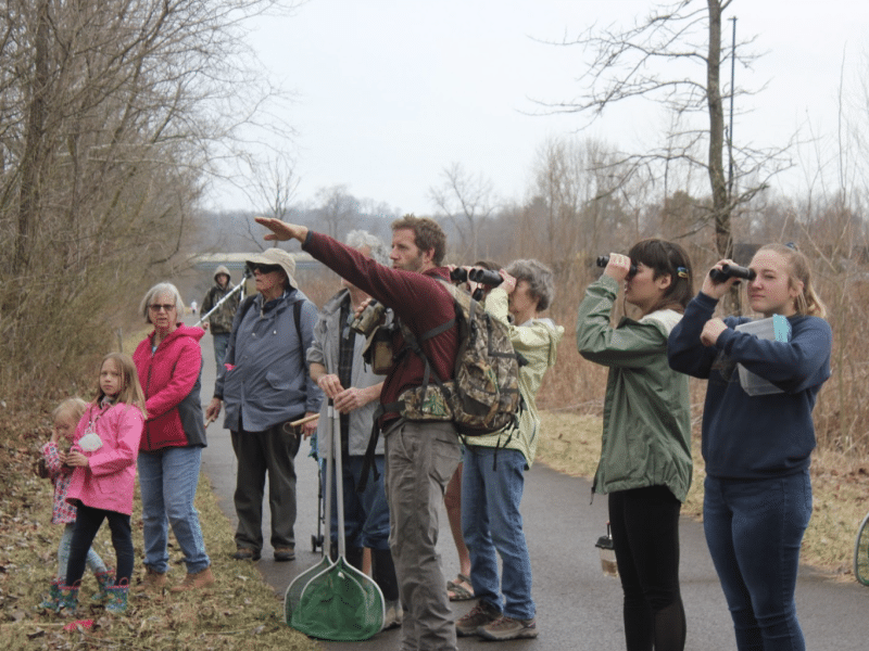 A group of birdwatchers, ranging from children to adults, looks into the distance through binoculars as a guide points out a bird.